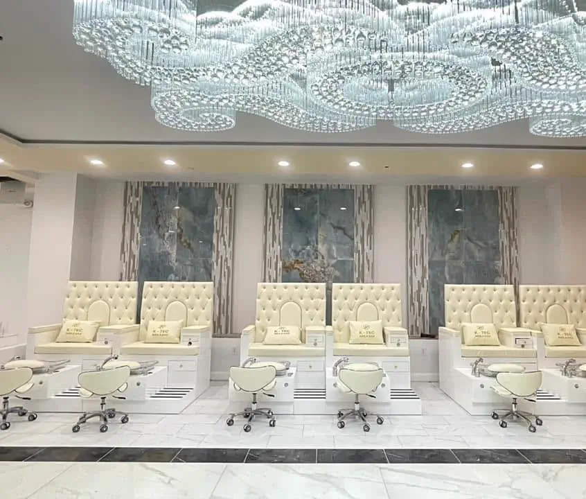 A row of white tufted pedicure chairs under a wide crystal chandelier, with marble walls